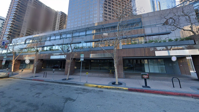 Street view of a modern multi-story office building with glass windows, trees along the sidewalk, and surrounding downtown buildings.