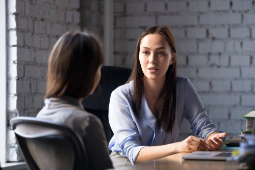 A professional woman in a light blue blouse is seated at a desk, attentively listening and speaking to another woman in a gray sweater. The setting is an office with a modern brick wall background.