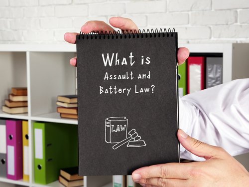 A person holding a notebook titled "What is Assault and Battery Law?" with legal icons on the cover, set against a background of office shelves.